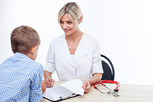 Attractive doctor is talking with a boy. They are sitting at the table and smiling. The woman is looking at the child happily and writing down some notes. Isolated on background